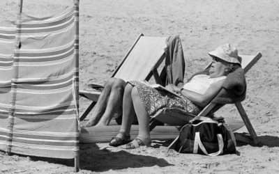 A woman holidaymaker wearing a floppy sun hat sleeps in a deck chair behind a windbreak on the sandy beach at Tenby, Pembrokeshire, South Wales. A man lies on an inflatable sunbed next to her. Only his legs can be seen     Date: 1988